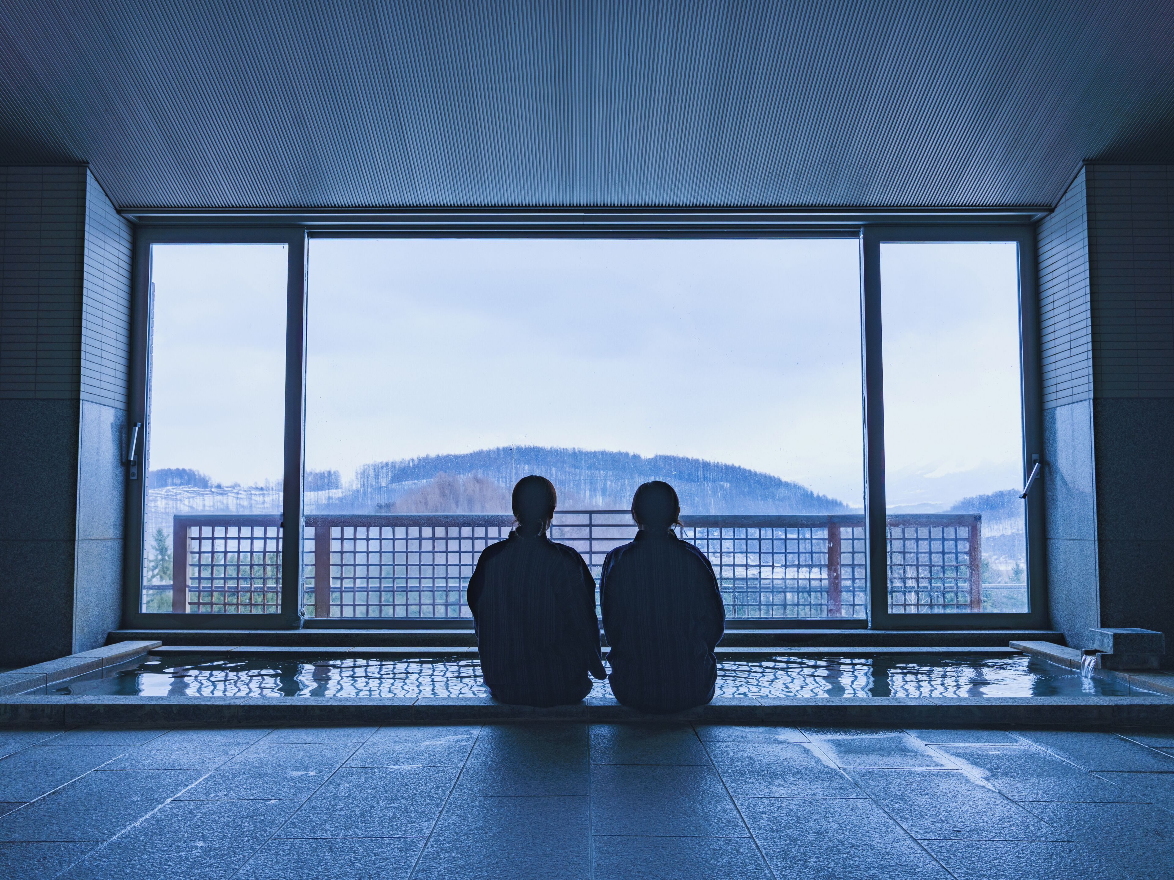 Two figures seated by a large window with a view of a snowy landscape.