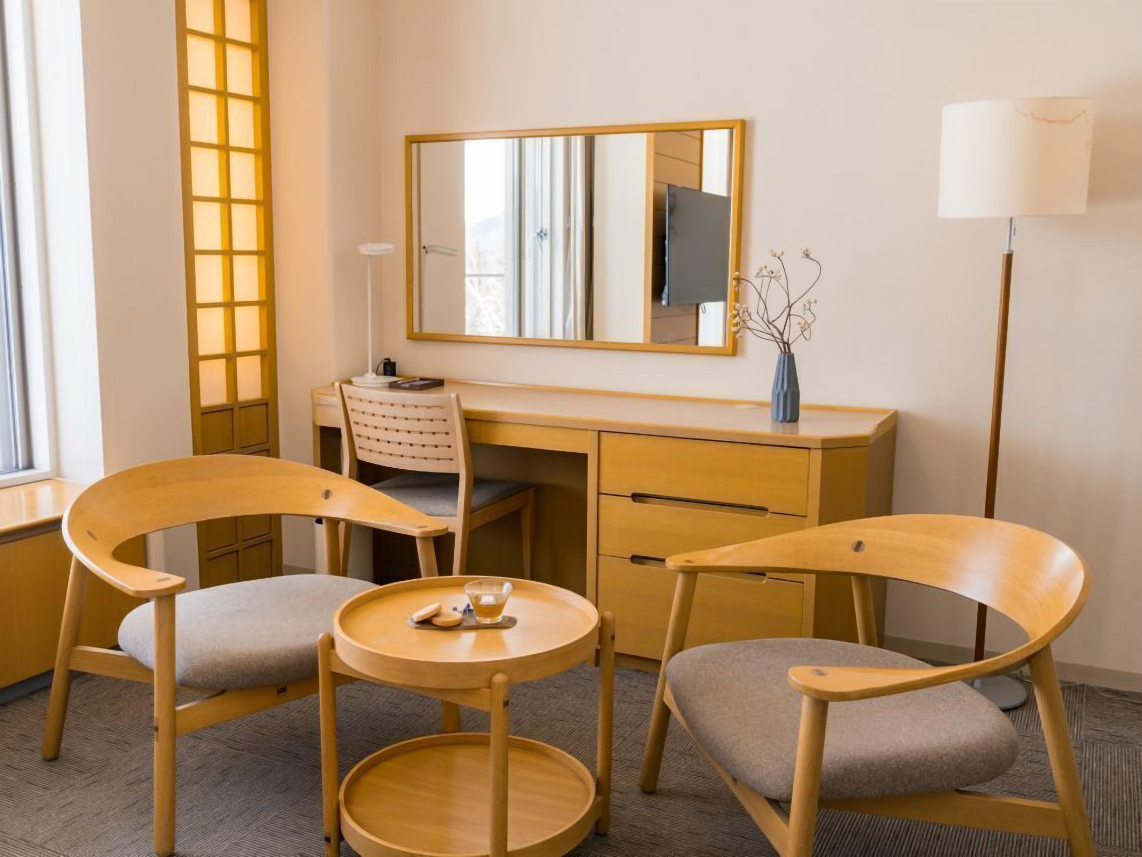 A hotel room with a desk, two chairs, a round table, a mirror, a lamp, and a vase with dried flowers.
