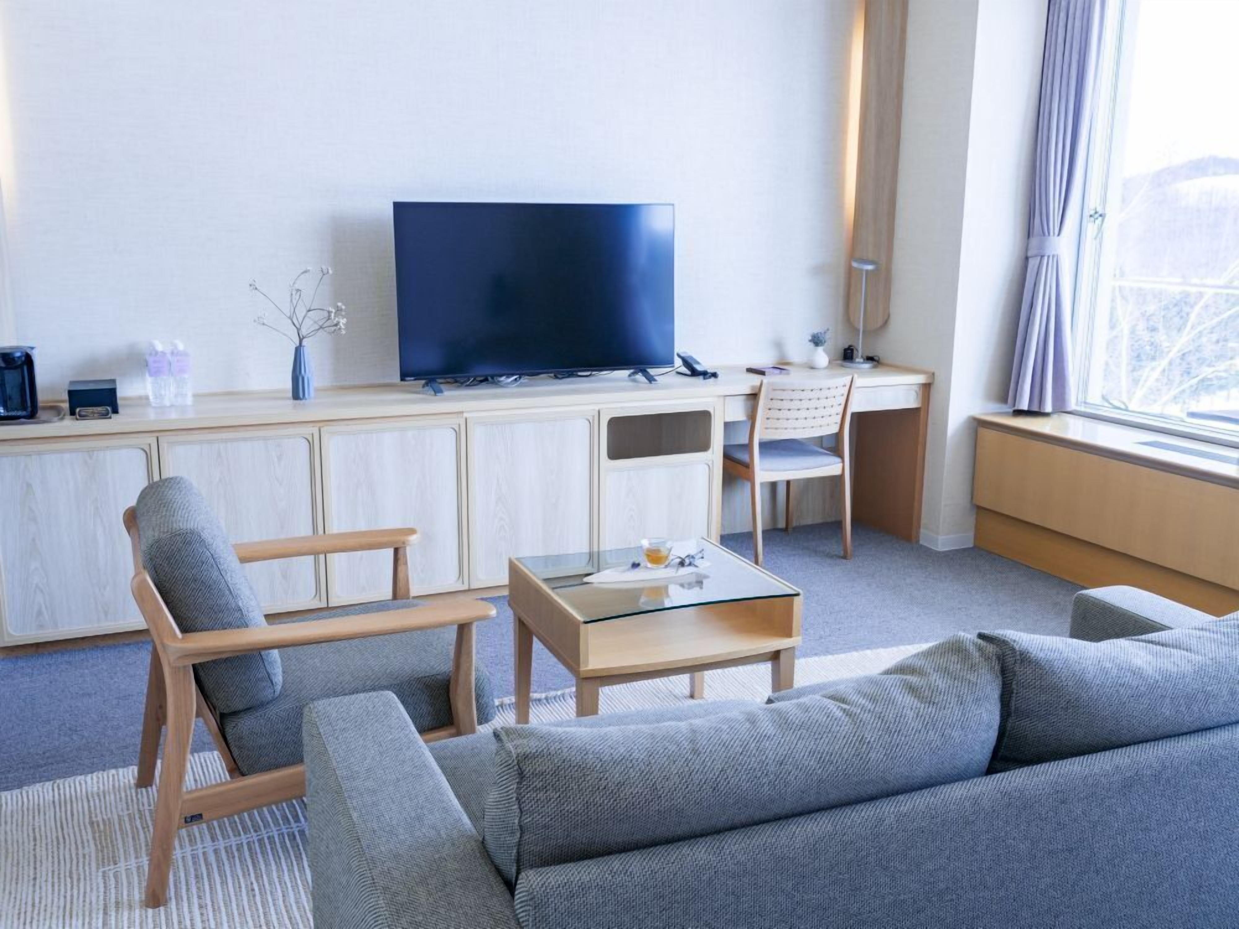 A modern hotel room with a flat-screen TV, a wooden desk, a glass coffee table, a sofa, and a chair.