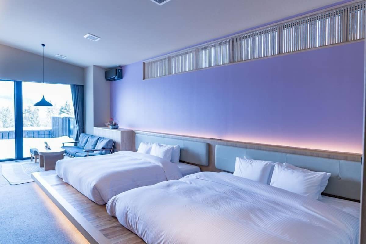 A modern hotel room with two large beds, a sofa, a small table, and a large window.