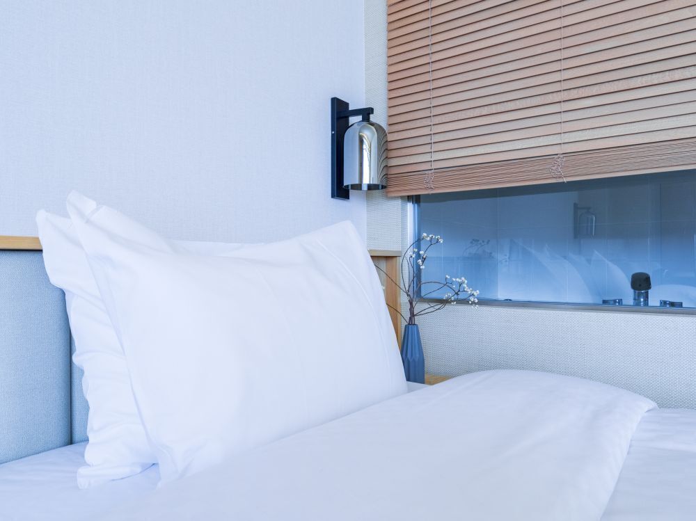 A bed with white bedding, a wall-mounted light fixture, and a window with blinds.