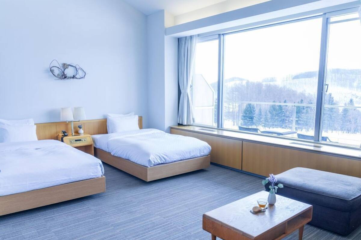 A hotel room with two beds, a wooden nightstand, a table, and a large window with a view of snow-covered trees.