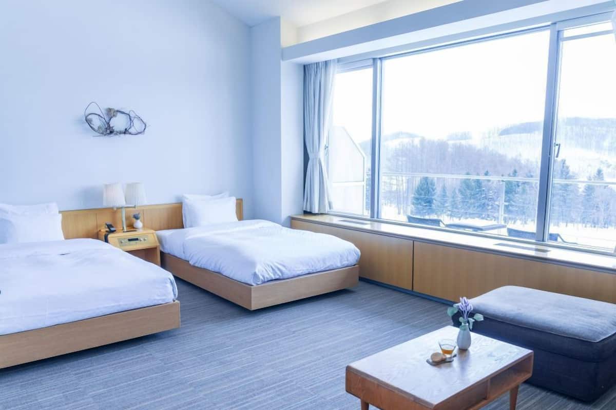 A hotel room with two beds, a wooden nightstand, a table, and a large window with a view of snow-covered trees.