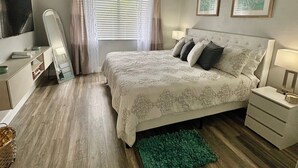 Une chambre à coucher avec un lit, un ventilateur de plafond, une table de chevet, une lampe, un miroir, une fenêtre avec des rideaux et un tapis vert.