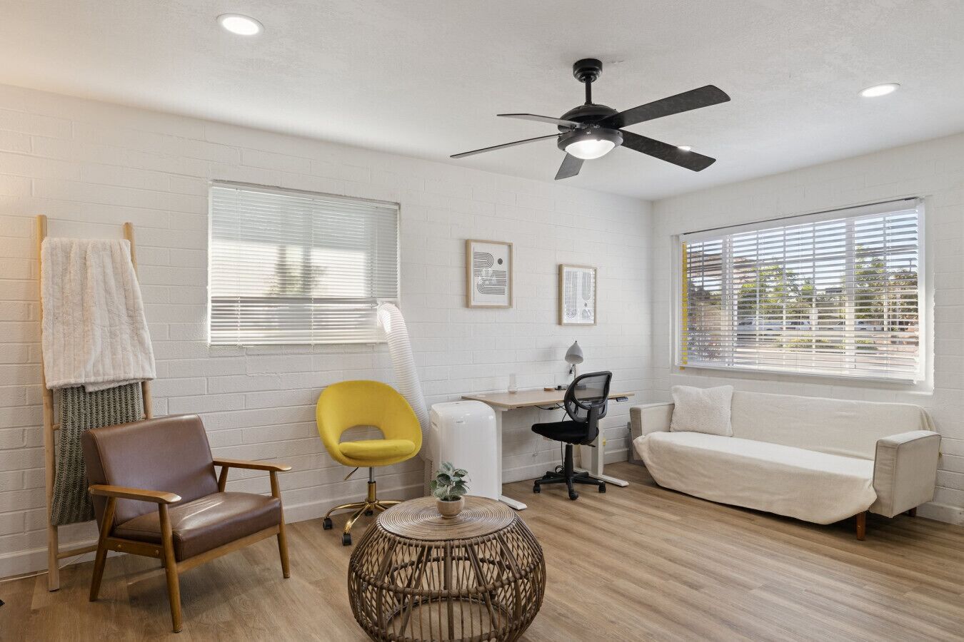 Una sala de estar moderna con un sofá, una butaca amarilla, un sillón de cuero marrón, una mesita redonda de mimbre, un escritorio con silla y un ventilador de techo.