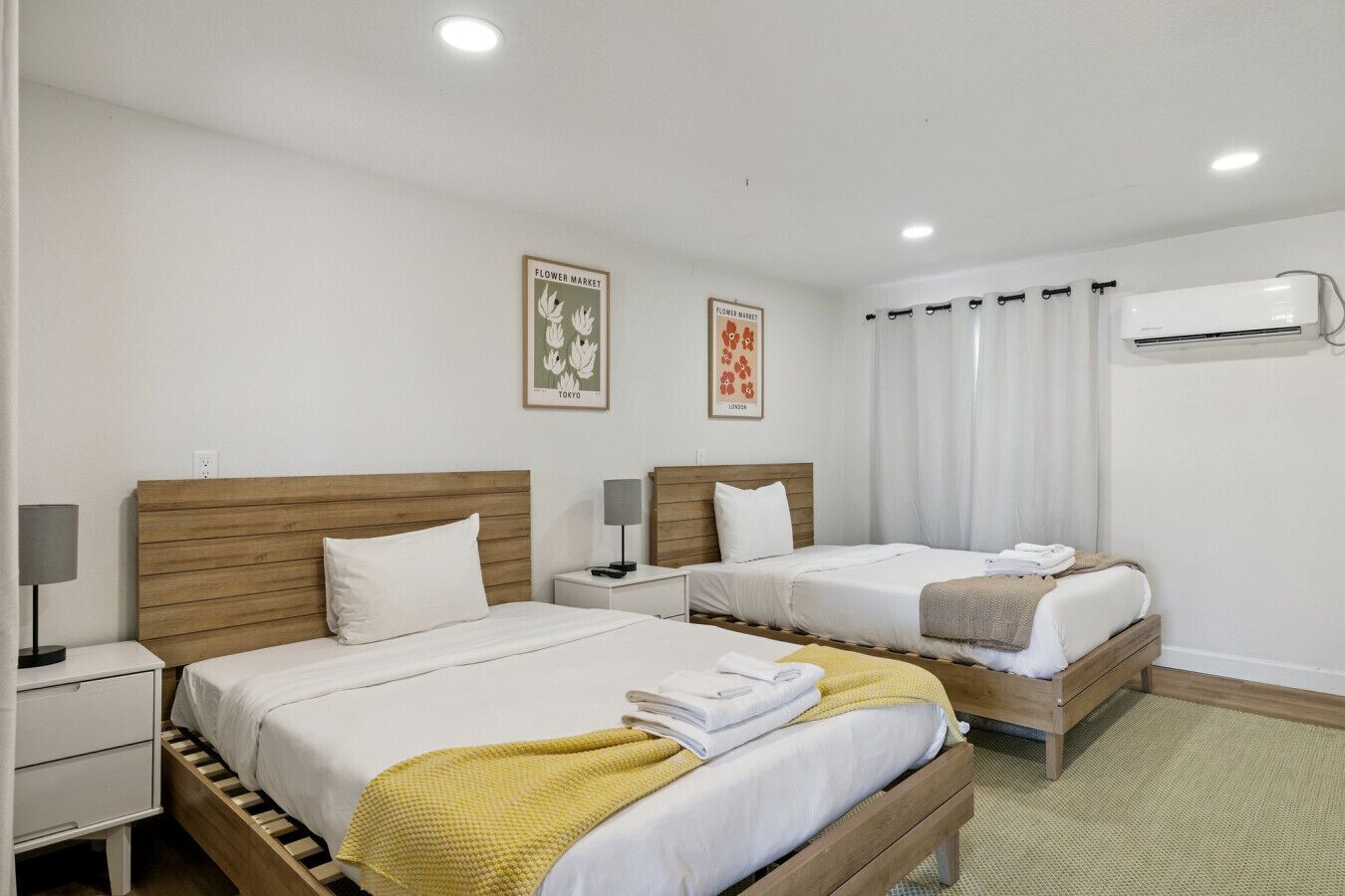 A hotel room with two beds, a bedside table with a lamp, a wall-mounted air conditioner, and two framed pictures on the wall.
