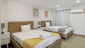 Une chambre d’hôtel avec deux lits, une table de chevet avec une lampe, un climatiseur fixé au mur et deux tableaux encadrés accrochés au mur.