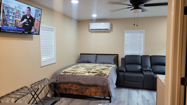 A bedroom with a bed, a black leather chair, an air conditioner, and a TV displaying a news program.