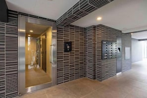 Un ascensor moderno con acabado metálico y puerta de vidrio, junto a una pared con diseño decorativo y una pantalla digital.