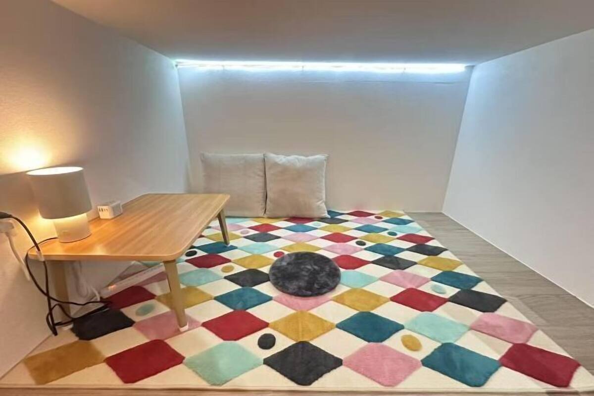 Ein kleiner, hell erleuchteter Raum mit einem farbenfrohen geometrischen Teppich, einem Holztisch mit Lampe und einem Bett mit Kissen.