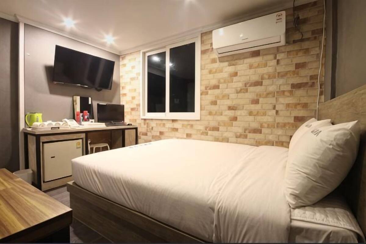 A bedroom with a bed, a desk with a computer, a TV, and an air conditioning unit.