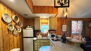 A compact kitchen with wooden cabinets, a refrigerator, a microwave, and a sink. There are woven baskets on the wall and a small dining area with a table and chairs. - The Apartment of GRACE! (Corpus Christi)
