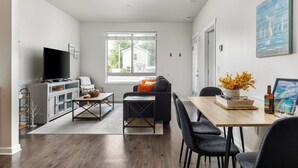Ein modernes Wohnzimmer mit einer Couch, einem Couchtisch und einem Fernseher auf einem Schrank. Ein Fenster bietet Blick auf ein Haus.
