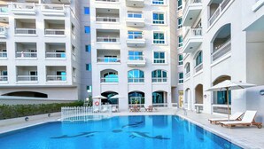 A swimming pool with lounge chairs and a lifebuoy, surrounded by a multi-story building with balconies and windows. - Relaxing ST Getaway Balcony, Pool & Gym Access (Dubai)