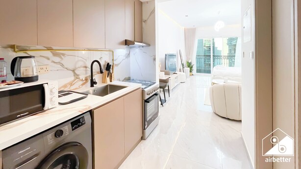 A modern kitchen with a washing machine, a microwave, and a sink. There is a dining area with a sofa and a TV. The room has a large window with a view of buildings. - Relaxing ST Getaway Balcony, Pool & Gym Access (Dubai)