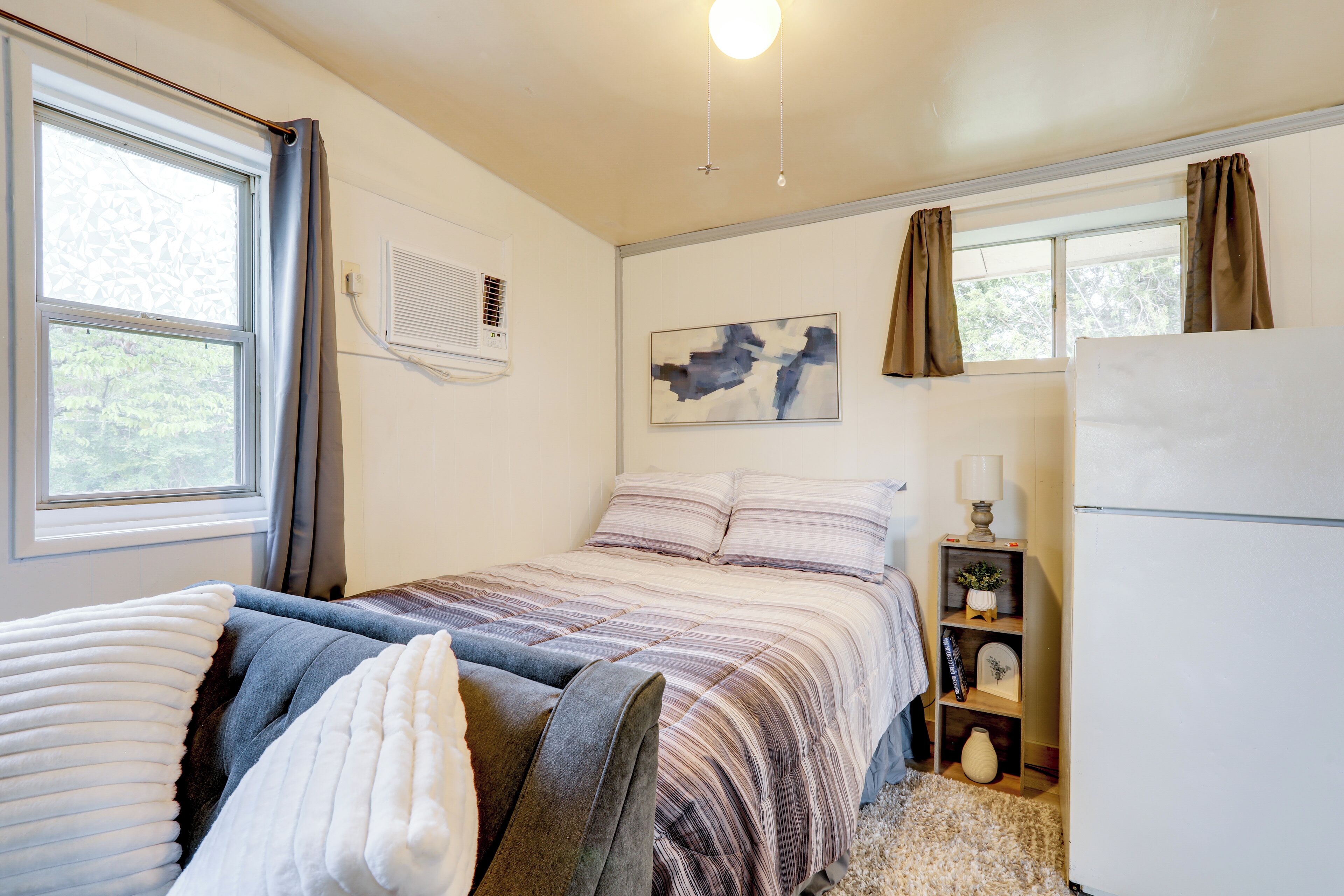 Een slaapkamer met een bed, nachtkastje, lamp, wandairconditioner, raam met gordijnen en een schilderij aan de muur.