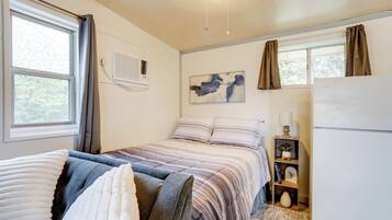 Een slaapkamer met een bed, nachtkastje, lamp, wandairconditioner, raam met gordijnen en een schilderij aan de muur.