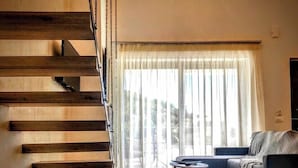 A modern living room with a wooden staircase, a chandelier, a sofa, and a view through large windows. - LefkoParga luxury Suites  (Parga)