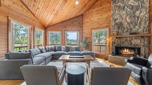 A cozy living room with a stone fireplace, wooden ceiling, and large windows offering a view of the outdoors.