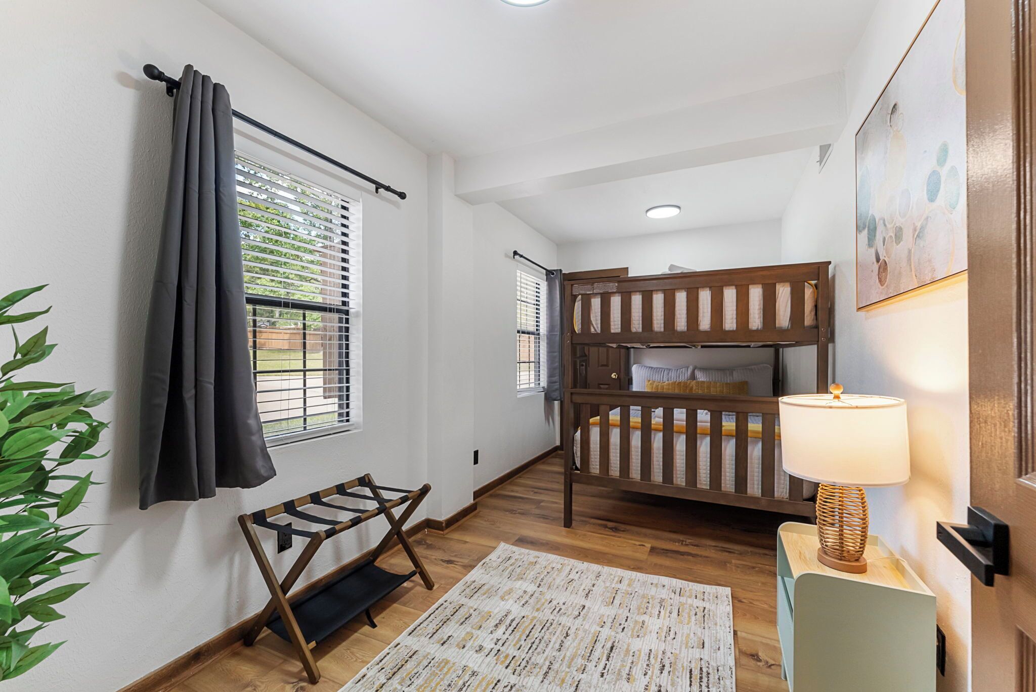 Une chambre avec un lit superposé, une table de chevet avec une lampe, un petit tapis, une plante et une fenêtre avec des stores.