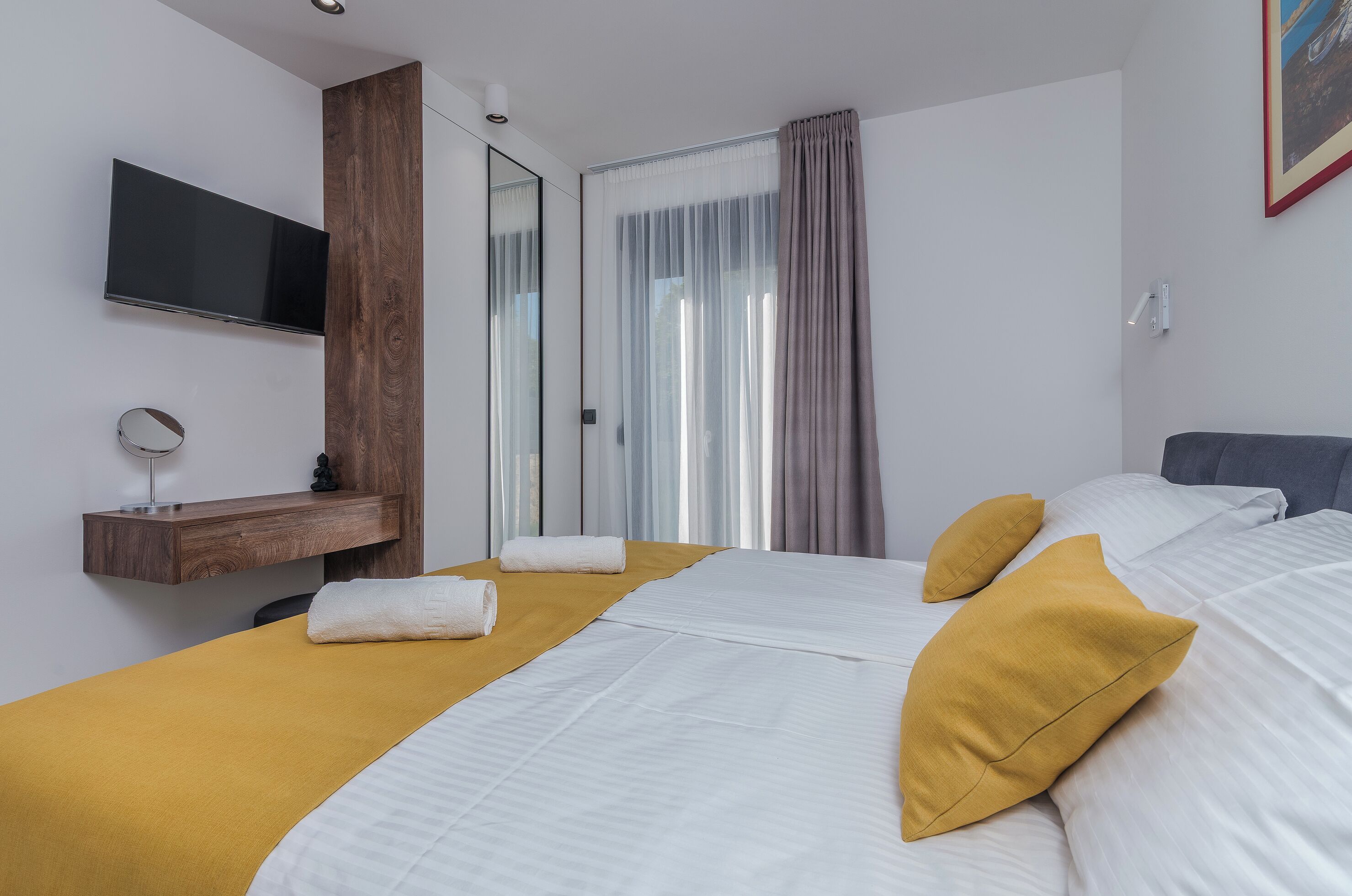 Une chambre d’hôtel avec un lit, un téléviseur fixé au mur, une étagère en bois avec un miroir et une fenêtre avec des rideaux.