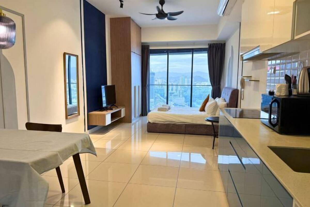 A modern hotel room with a large bed, a dining table, a kitchenette, and a city view.