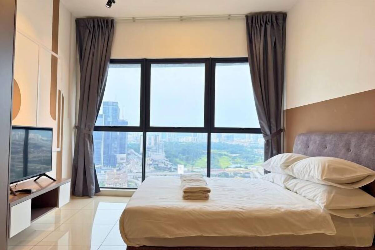 A modern hotel room with a large bed, a flat-screen TV, and a city view through large windows.
