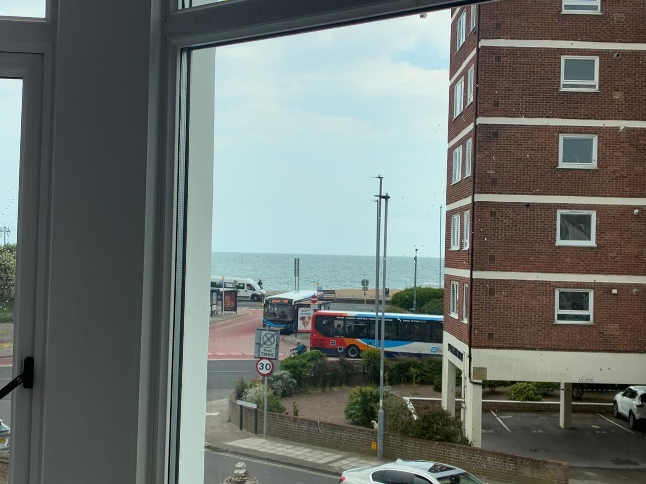 A view from a window overlooking a street with buses, a parking area, and a building with a brick facade.