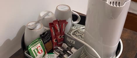 Eine Kaffeemaschine, eine Tasse, ein Löffel, eine Kaffeepackung, eine Teepackung, eine Zuckerpackung, eine Milchpackung und eine Kaffeeweißerpackung auf einem Tablett.