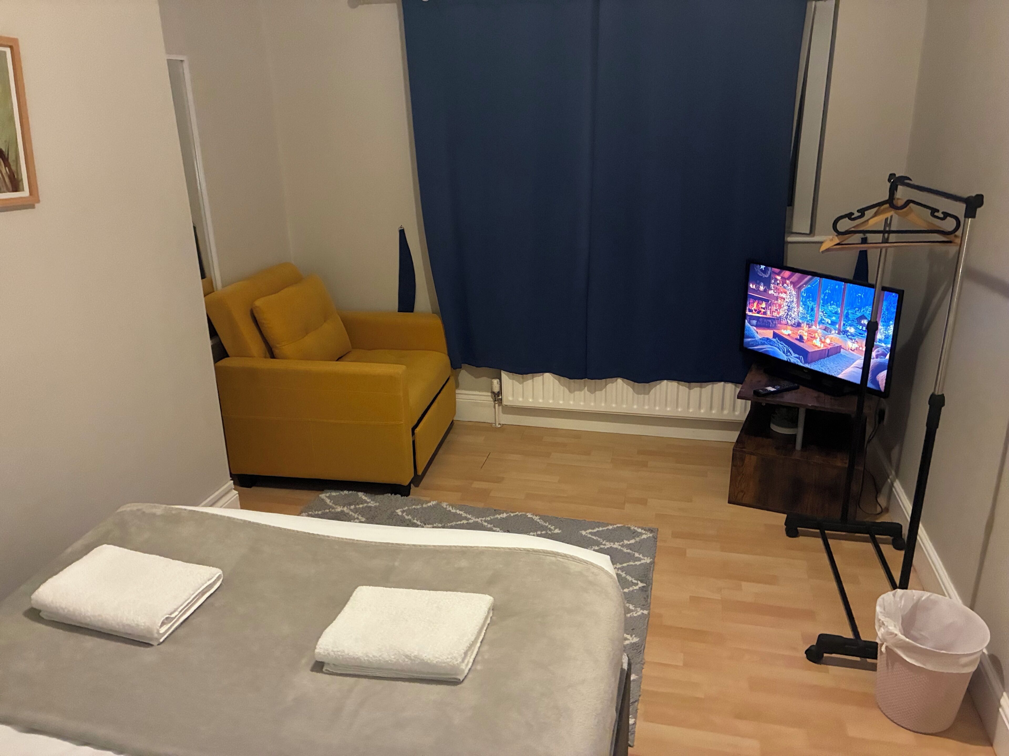 A hotel room with a bed, a yellow armchair, a television, and a window with a blue curtain.