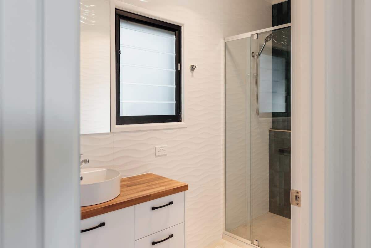 A modern bathroom with a glass-enclosed shower, a wooden vanity with a white sink, and a window above the mirror.