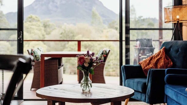 Una habitación con una mesa redonda de madera, un florero con flores, un sofá azul y vistas a la montaña a través de amplias ventanas.