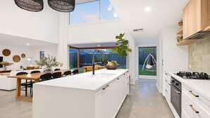 Eine moderne Küche mit einer zentralen Kochinsel, einem Essbereich mit Holztisch und Blick auf einen Garten durch große Fenster.