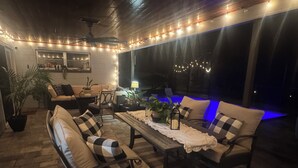 Un espacio al aire libre acogedor con mesa de madera, sillas con cojines e iluminación con luces colgantes.