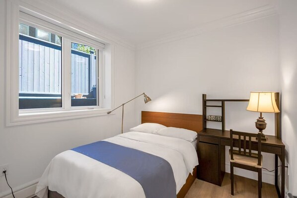 A bedroom with a bed, bedside table, lamp, and a window with a view of a blue fence. - Carol’s garden home in the best community (Vancouver)