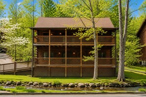 A two-story log cabin with a balcony, surrounded by a well-maintained lawn and a small stream. - EV Charger l Hot Tub l Fireplace l Arcade l BBQ (Pigeon Forge)