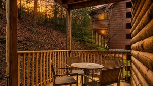 Eine Holzterrasse mit einem runden Tisch und Stühlen, mit Blick auf eine bewaldete Gegend, im Hintergrund ein Häuschen.