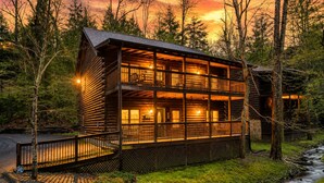 Eine zweistöckige Holzblockhütte mit Balkon, umgeben von Bäumen und einem Bach.