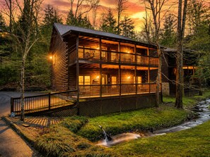 Eine zweistöckige Holzblockhütte mit Balkon, umgeben von Bäumen und einem Bach.