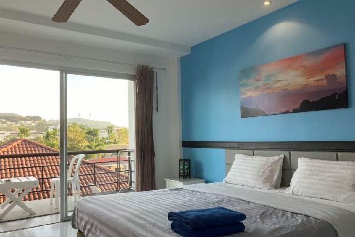 A hotel room with a bed, a painting, a nightstand, and a view of the city.