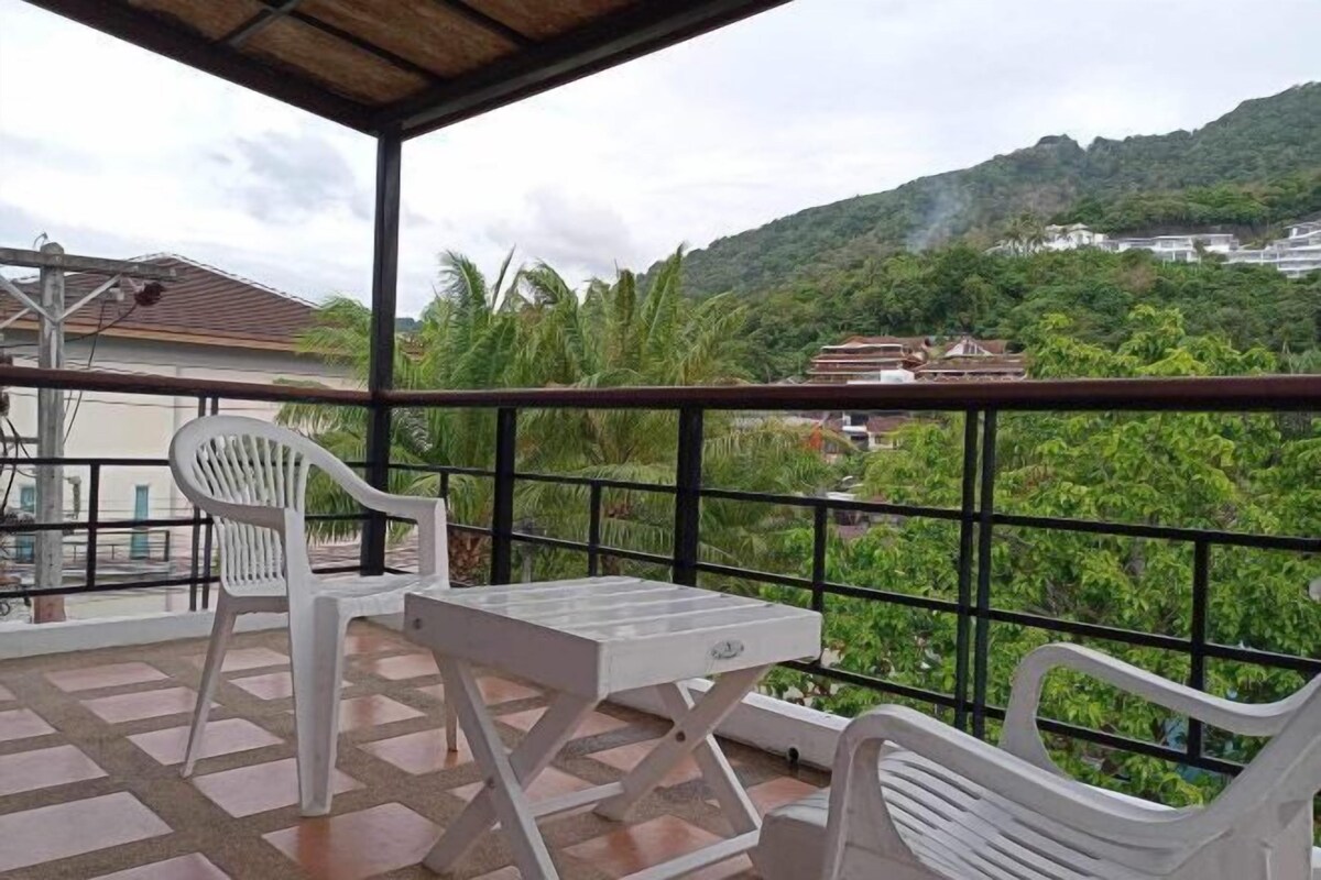 A covered balcony with a table and two chairs, overlooking a lush green hillside with buildings in the background.