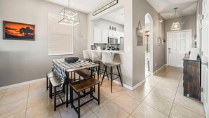 Una cocina moderna con armarios blancos, una zona de comedor con mantel a cuadros, y vista al área de estar a través de un arco.