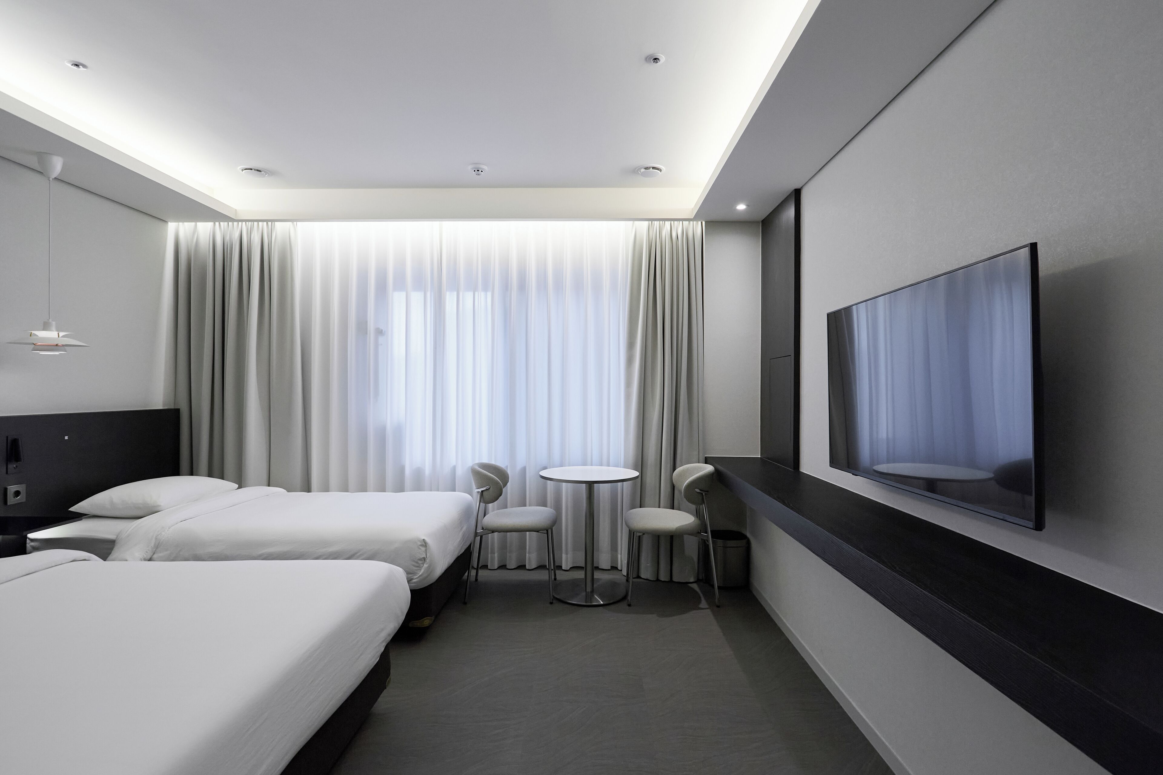 A hotel room with two beds, a small round table, two chairs, a TV on the wall, and a large window with curtains.