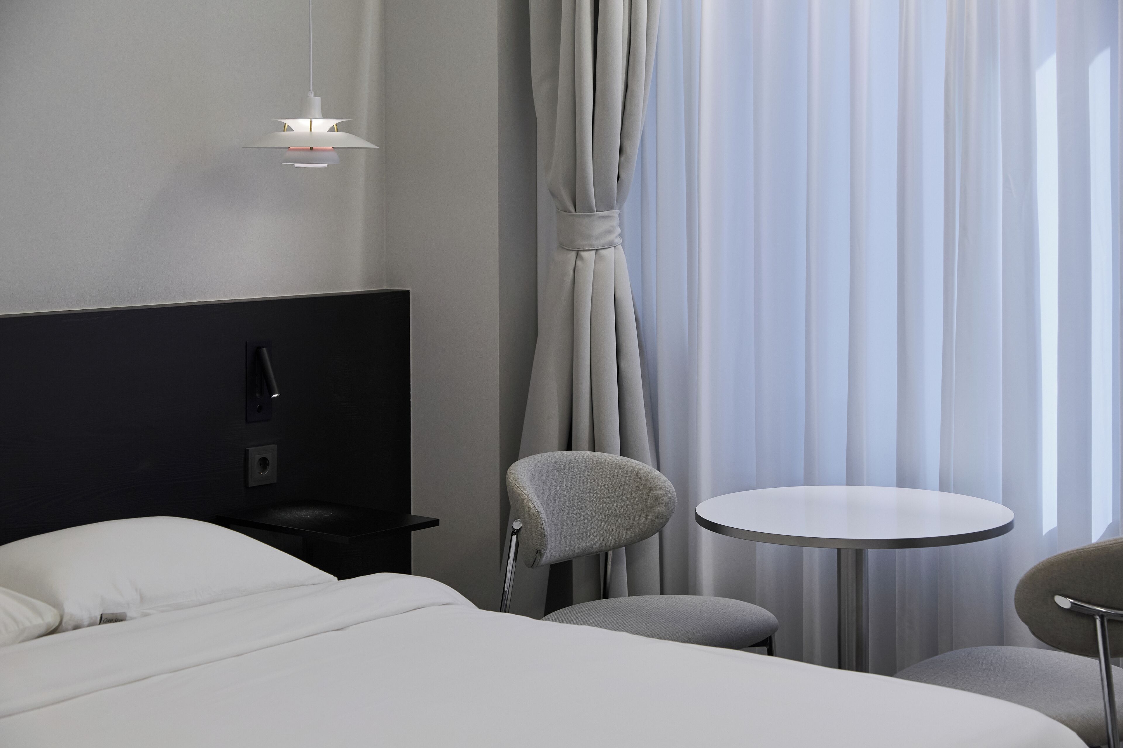 A modern hotel room with a bed, a small table, a chair, and a hanging light fixture.