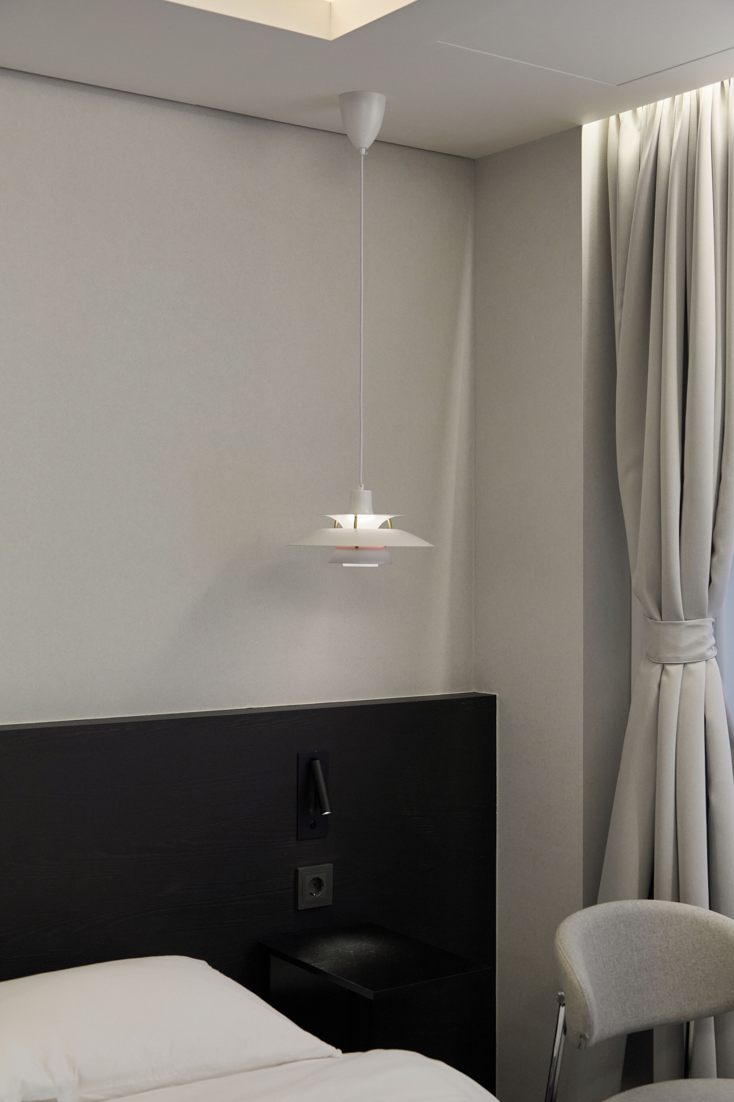 A modern hotel room with a bed, a desk, a chair, a hanging light fixture, and a window with curtains.