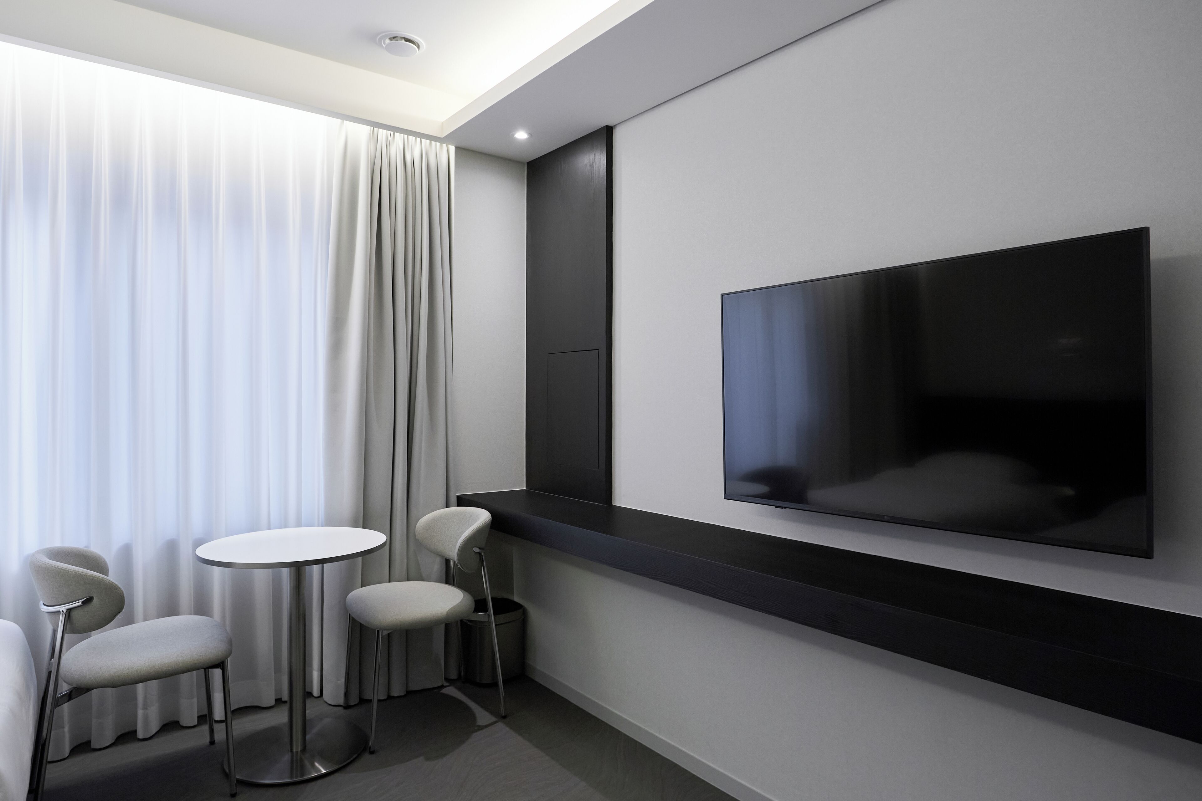 A modern hotel room with a large flat-screen TV mounted on the wall, a round table with two chairs, and a window with sheer curtains.