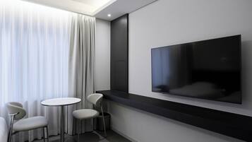 A modern hotel room with a large flat-screen TV mounted on the wall, a round table with two chairs, and a window with sheer curtains.
