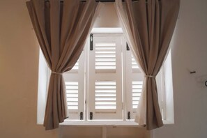 A window with beige curtains, white shutters, and a view of a white wall. - Carnarvon 73 (George Town)