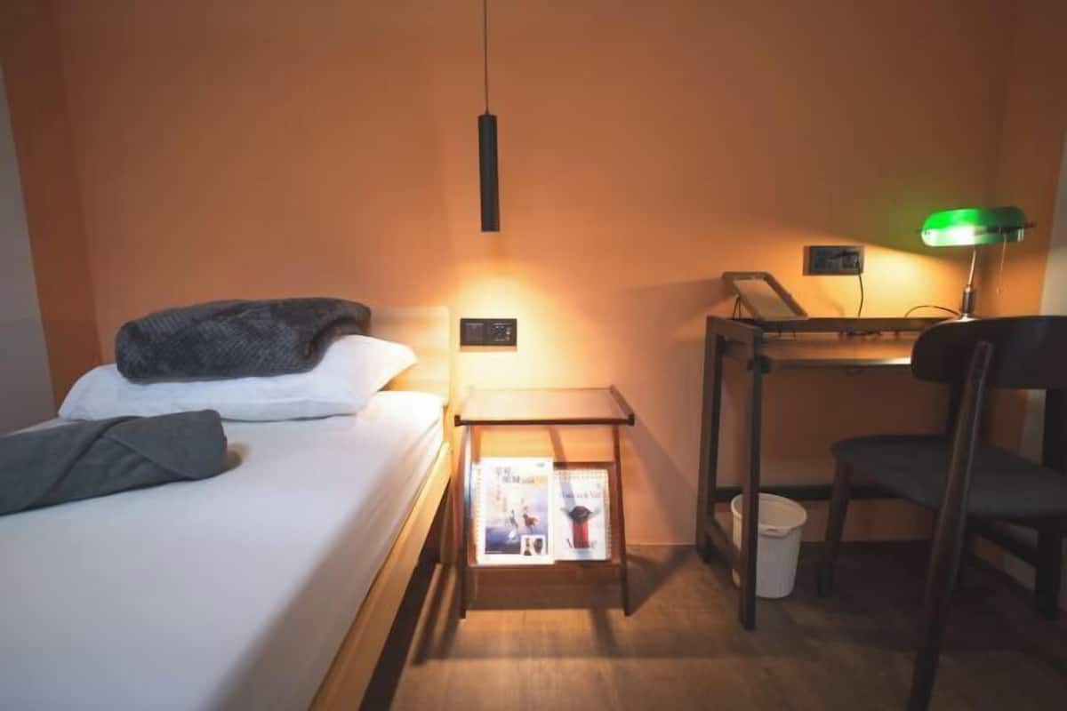 A hotel room with a bed, a desk, a chair, a nightstand with a lamp, and a small trash bin.