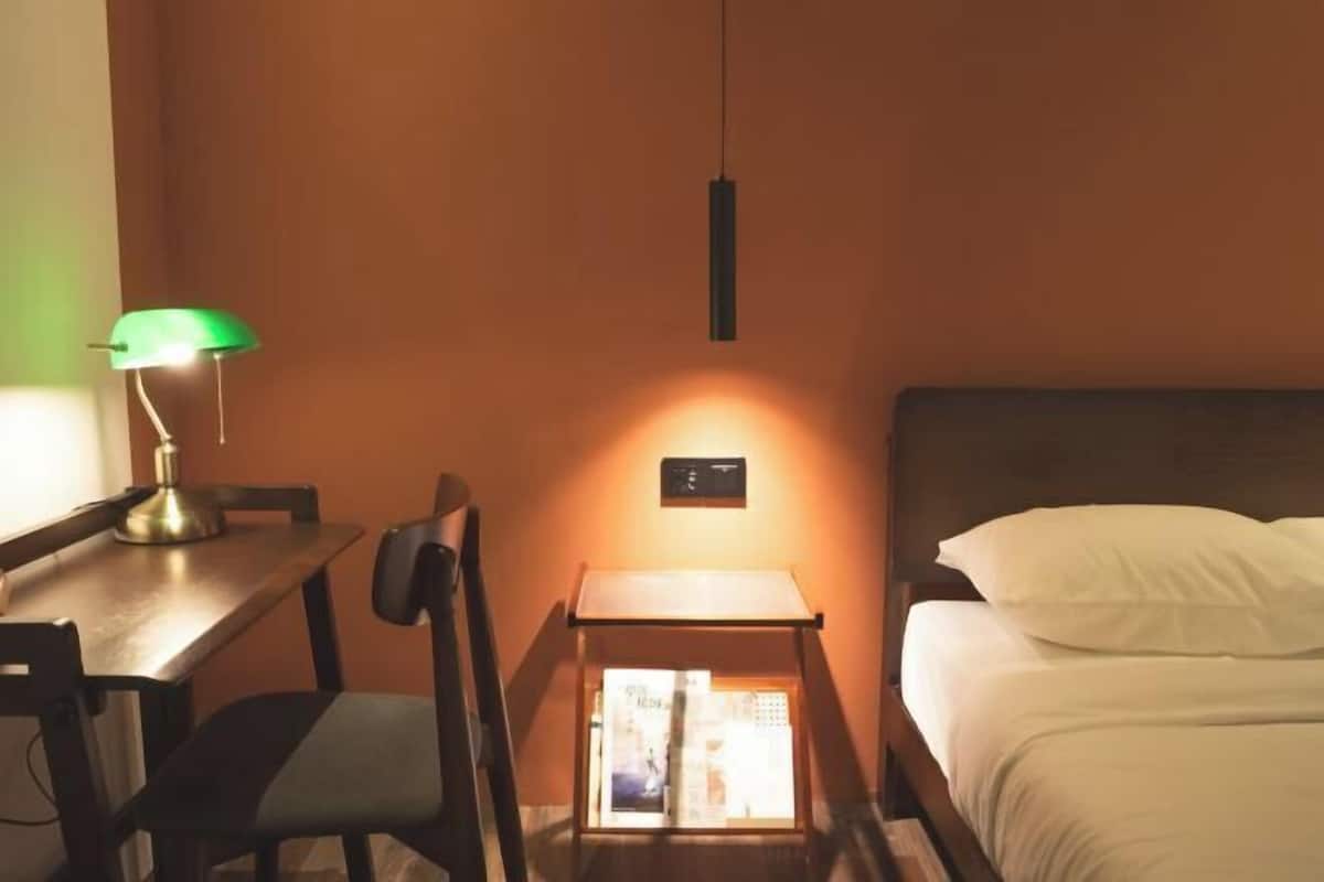 A hotel room with a bed, a desk with a lamp, a chair, a small table with a book, and a wall-mounted light fixture.